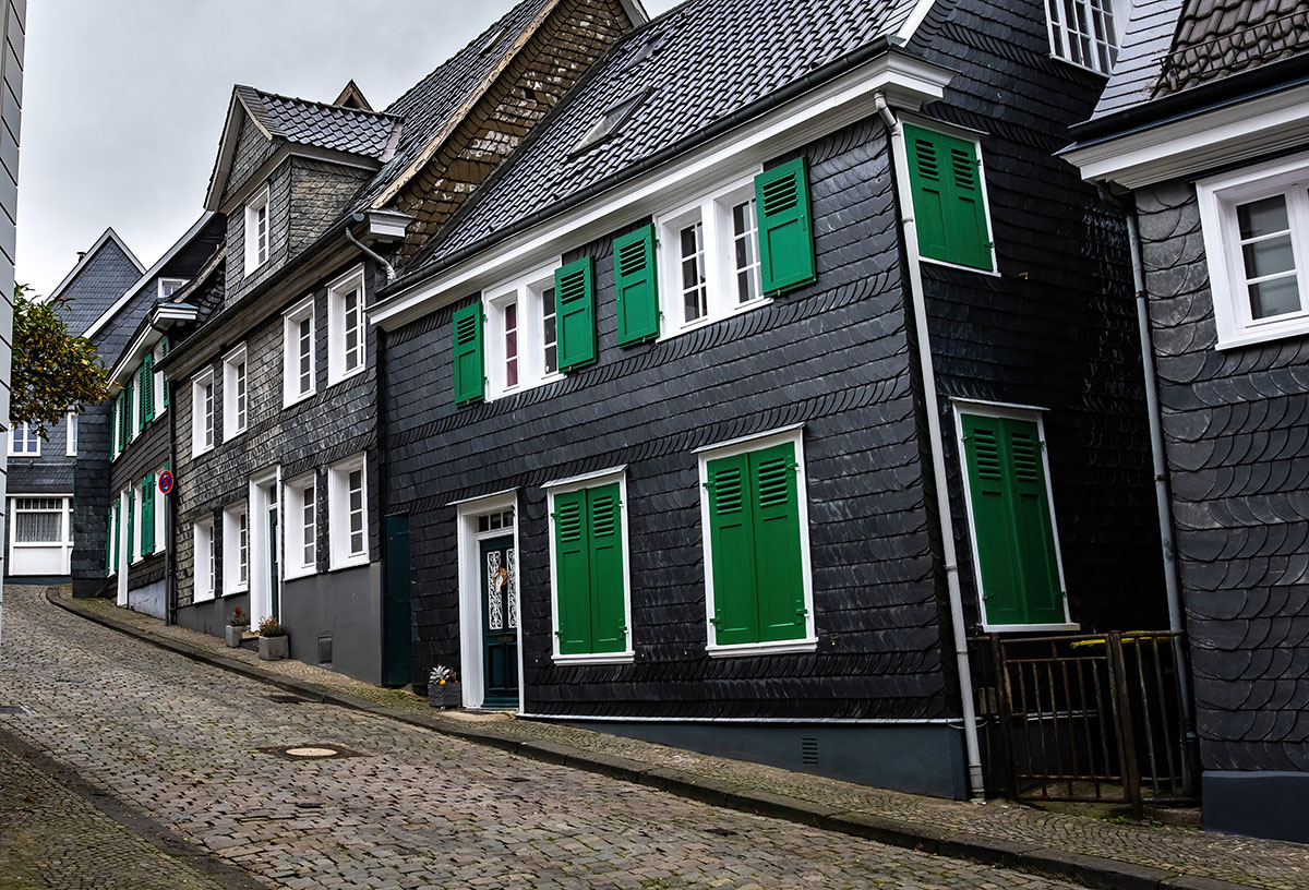 Traditionelle Häuser mit Schieferfassaden und grünen Fensterläden in einer gepflasterten Straße, die sich durch eine historische Altstadt schlängelt.