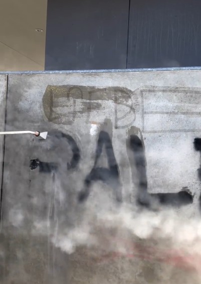 Fassadenreinigung mit Niederdrucktechnik an einer Betonwand in Mannheim: Entfernen von schwarzer Graffiti-Farbe mithilfe eines Heißwasserstrahls.