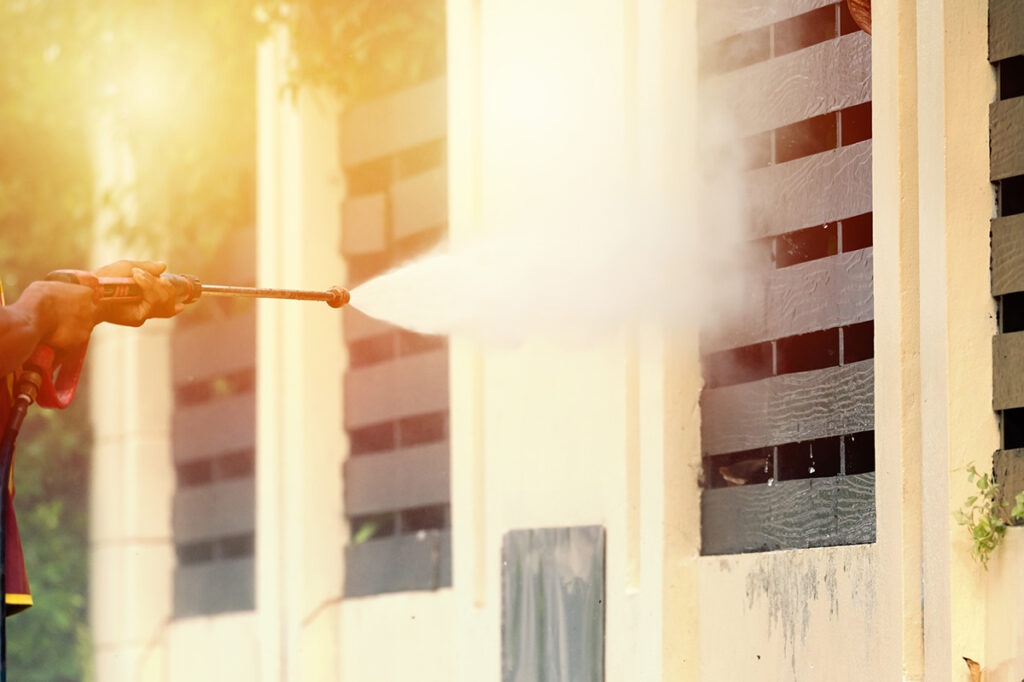 Person sprüht mit einem Hochdruckreiniger gegen eine helle Fassade mit Lamellenfenstern bei Sonnenlicht.