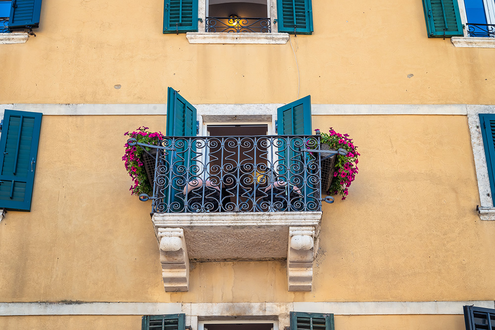 Balkonfassade mit gelbem Putz, grünen Fensterläden und Blumenkästen an einem Mehrfamilienhaus.