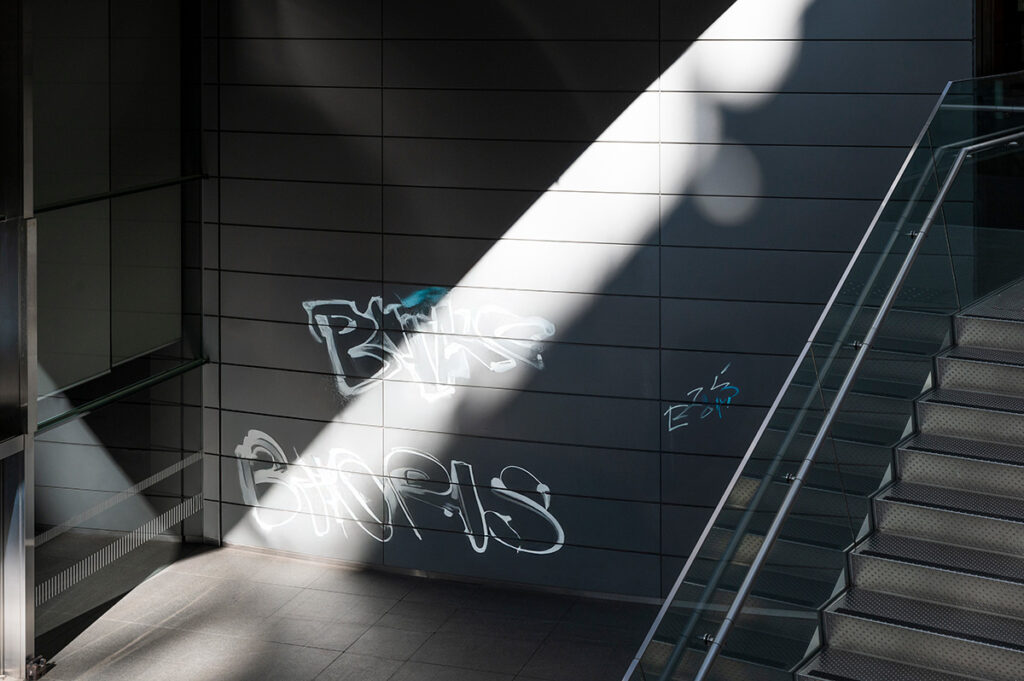 Graffiti auf dunkler Metallfassade in einem modernen Treppenhaus mit Lichteinfall.
