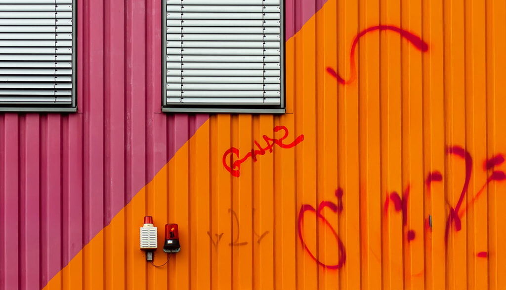 Graffiti auf einer farbigen Metallfassade mit orange-roten und pinken Paneelen.