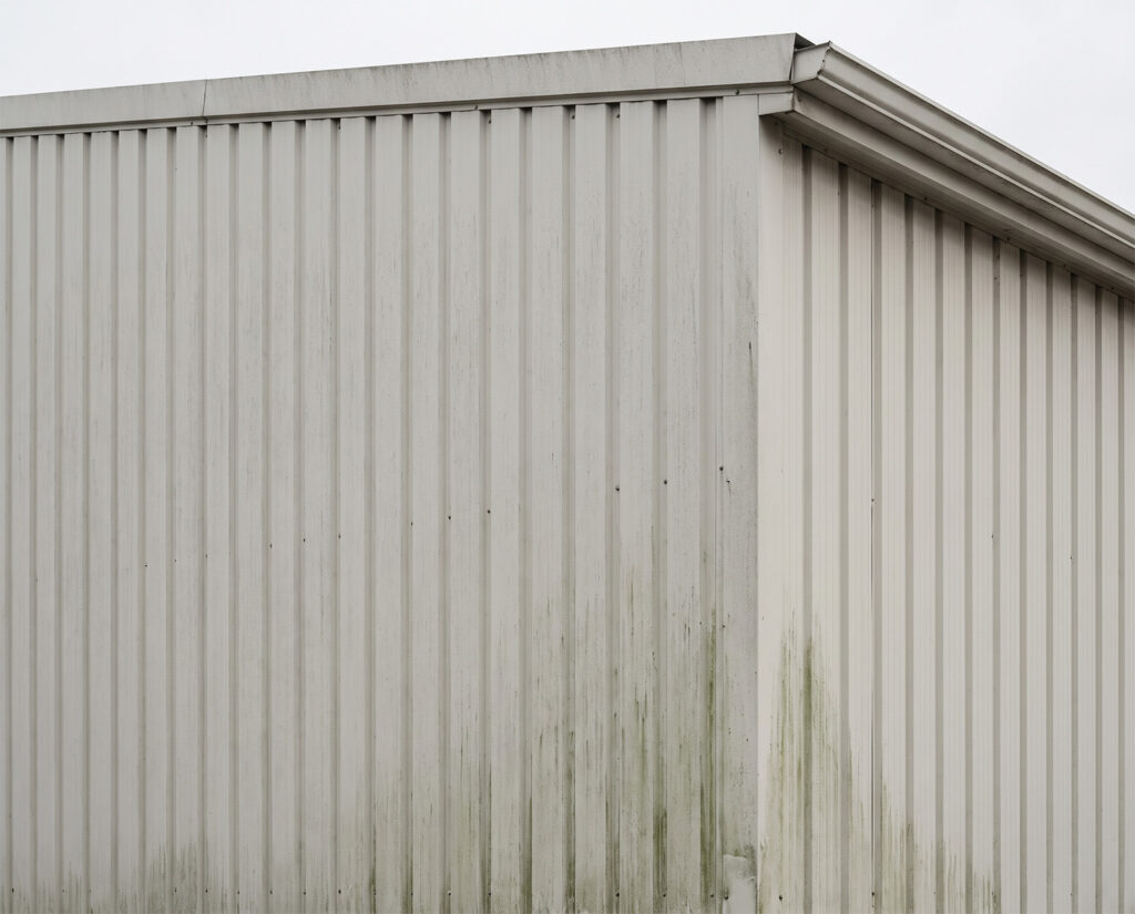 Hallenfassaden reinigen: helle Trapezblech-Fassade einer Halle mit grünlichen Algen- und Schmutzstreifen im unteren Bereich.